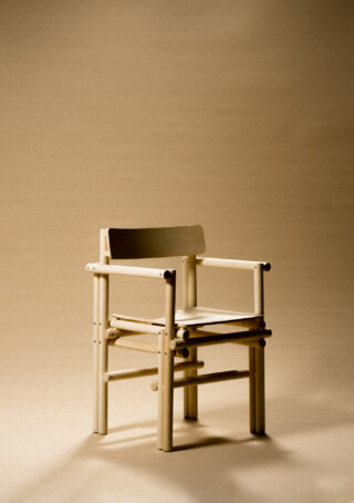 Auf einem schlichten, sanft beleuchteten beigen Hintergrund steht ein Holzstuhl mit Armlehnen und Lattenrückenlehne.