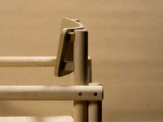 Nahaufnahme eines Holzstuhls mit geschwungener Rückenlehne und sichtbaren Schrauben vor beigem Hintergrund.