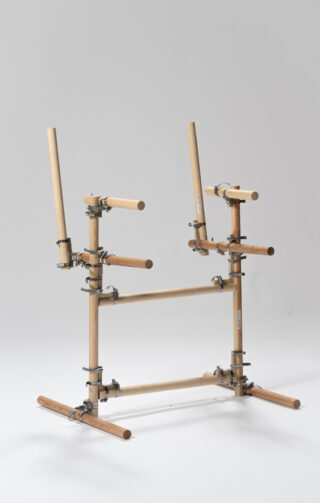 Eine aus zylindrischen Stäben und Metallklammern zusammengesetzte Holzkonstruktion, die auf einer weißen Fläche vor einem neutralen Hintergrund steht.