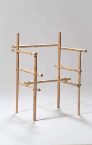 Eine minimalistische, mit Dübeln und Bolzen zusammengefügte Holzkonstruktion, bestehend aus vertikalen und horizontalen Stäben, steht auf einem weißen Untergrund.