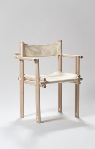 Ein minimalistischer Stuhl mit einem hellen Holzrahmen und einer Sitzfläche und Rückenlehne aus cremefarbenem Segeltuch, fotografiert vor einem schlichten grauen Hintergrund.