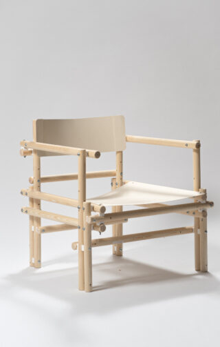 Ein minimalistischer Holzstuhl mit hellem Holzrahmen, Sitz und Rückenlehne aus beigem Stoff und sichtbaren Metallverschlüssen auf schlichtem Hintergrund.