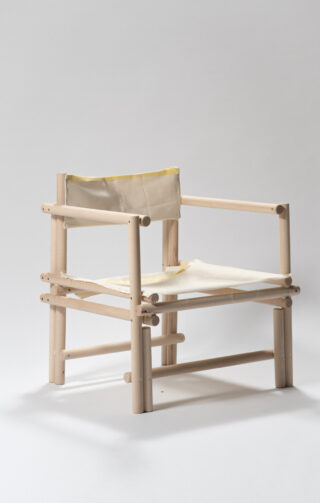 Ein minimalistischer Stuhl mit hellem Holzrahmen und Sitzfläche und Rückenlehne aus cremefarbenem Segeltuch vor einem schlichten hellen Hintergrund.