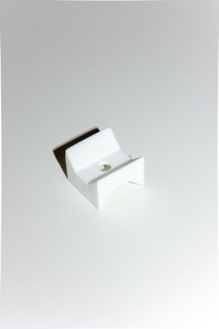 Eine kleine weiße Kunststoffhalterung mit einem Loch in der Mitte sitzt auf einer schlichten weißen Oberfläche und wirft einen Schatten nach links.