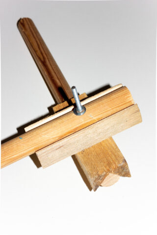 Ein selbstgemachtes Holzwerkzeug mit einem vertikalen Teil, das mit einer Schraube und einer Flügelmutter an einer horizontalen Basis befestigt ist, fotografiert vor einem schlichten weißen Hintergrund.