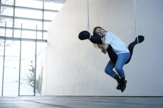 Eine Person in Jeans und weißem Hemd sitzt lächelnd auf einer schwebenden abstrakten schwarzen Skulptur und umarmt sie in einem modernen, lichtdurchfluteten Raum.