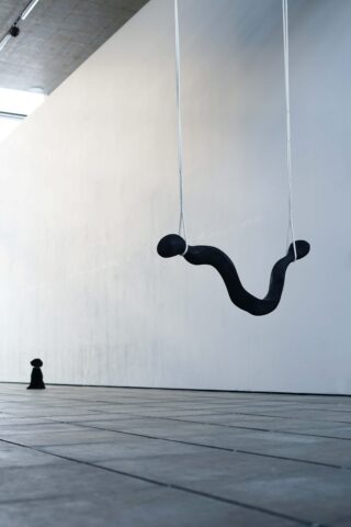 In einem minimalistischen Galerieraum hängt ein gebogenes, schwarzes abstraktes Objekt an zwei Seilen von der Decke; im Hintergrund steht auf einem gefliesten Boden eine kleine schwarze Skulptur.