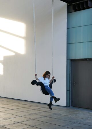 Eine Person in Jeans und Stiefeln sitzt in einem Innenraum mit gefliestem Boden und geschlossener Tür auf einer schwarzen Schaukel, die von der Decke hängt.
