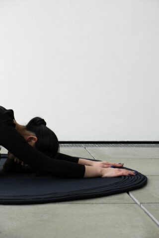 Person in schwarzer Kleidung übt eine Vorwärtsdehnung oder die Stellung des Kindes auf einer dunklen Matte, die auf einem gefliesten Boden vor einer schlichten weißen Wand steht.