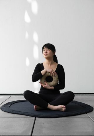 Eine Person in schwarzer Kleidung sitzt im Schneidersitz auf einer schwarzen Matte und hält ein rundes Kissen vor einer schlichten weißen Wand.