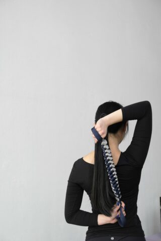 Eine Person mit langen schwarzen Haaren verwendet einen geflochtenen Übungs-Stretchgurt hinter ihrem Rücken und zieht mit einem Arm, während sie von der Kamera wegblickt.