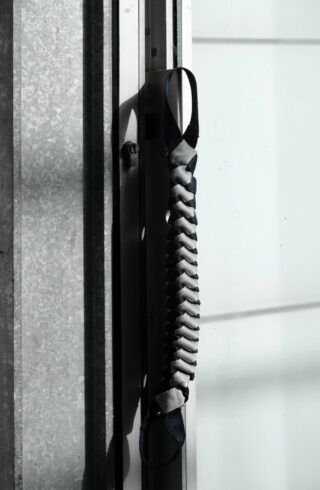 Ein geflochtener Riemen mit Schlaufen an beiden Enden hängt an einem Metallrahmen in der Nähe eines Fensters mit Milchglas.