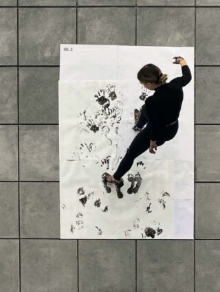 Eine Person in schwarzer Kleidung tritt auf große weiße Laken mit schwarzer Farbe und hinterlässt Hand- und Fußabdrücke auf einem gefliesten Boden.