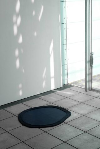 Eine dunkle, unregelmäßig geformte Matte liegt auf einem gefliesten Boden in der Nähe einer Glastür, und Sonnenlicht wirft Schatten an die Wand.