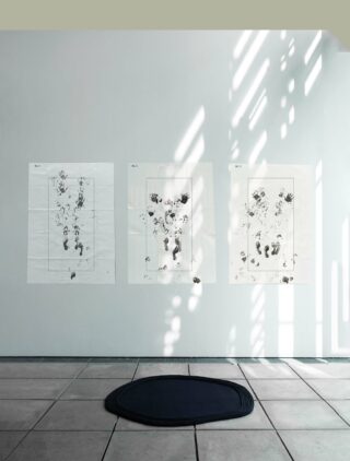 Drei große Blätter Papier mit abstrakten Hand- und Fußabdruckmotiven sind an einer weißen Wand über einer schwarzen runden Matte auf einem gefliesten Boden angebracht.