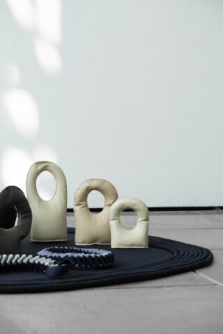 Mehrere kleine, kissenartige Objekte mit runden Griffen liegen auf einer runden schwarzen Matte neben einer geflochtenen Kordel vor einer hellen Wand und einem gefliesten Boden.