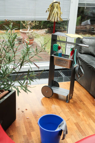 Ein Putzwagen mit Zubehör steht auf einem Holzboden neben einem blauen Eimer und einer Topfpflanze in einem hellen Raum mit großen Fenstern.
