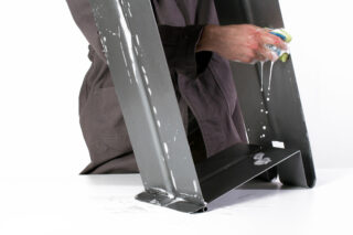 Eine Person in grauer Kleidung reinigt einen schwarzen Metallgegenstand mit einem Schwamm und Seifenwasser, wodurch Schaum und Tropfen auf einer weißen Oberfläche entstehen.