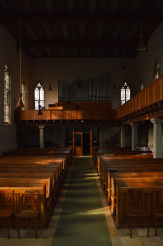 Sonnenlicht strömt in einen leeren Kirchenraum mit Holzbänken, einem grünen Mittelgangläufer, Buntglasfenstern und einer Orgel im hinteren Teil.