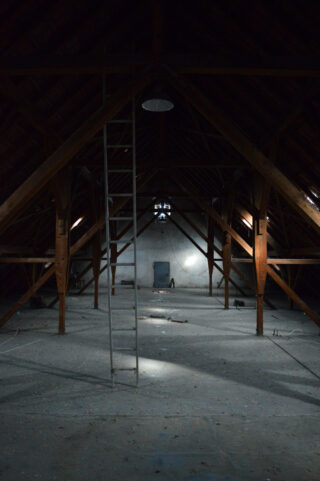 Ein schwach beleuchteter Dachboden mit Holzbalken, einer senkrechten Leiter, verstreutem Schutt und einem einzelnen Stuhl in der Nähe der Rückwand.