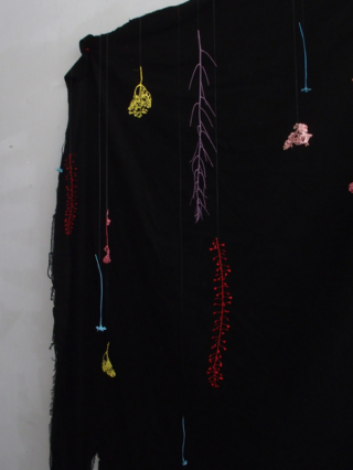 Bunte Häkelformen hängen an Schnüren vor einem drapierten schwarzen Stoffhintergrund vor einer weißen Wand.