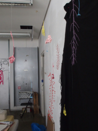 Ein schwach beleuchteter Raum mit bunten Drahtskulpturen, die von der Decke und der Wand hängen, einem schwarzen Vorhang und verschiedenen, überall verstreuten Gegenständen.