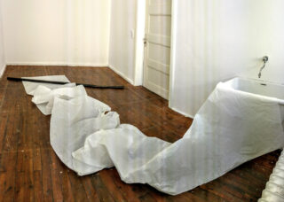 Ein weißes Laken oder Stoff ist auf einem Holzboden in einem ansonsten leeren Raum mit einer weißen Tür, einem an der Wand montierten Waschbecken und einem Heizkörper ausgebreitet.