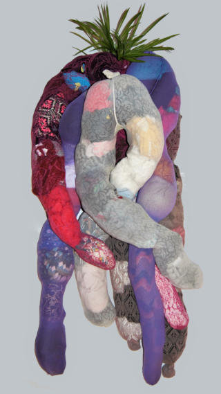 Eine weiche Skulptur aus bunten, gemusterten Stoffröhren ist vertikal angeordnet, mit einer grünen Pflanze an der Spitze, vor einem schlichten hellen Hintergrund.