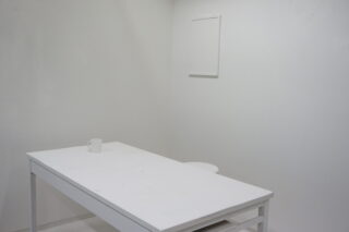 Ein schlichter weißer Raum mit einem weißen Tisch, einem weißen Stuhl und einer einzelnen weißen Tasse auf dem Tisch. Wände und Boden sind ebenfalls weiß und schaffen so ein minimalistisches Ambiente.