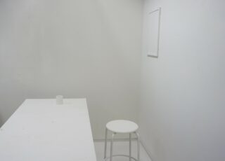 Ein kleiner, schlichter weißer Raum mit einem weißen Tisch, einer einzelnen Rolle Toilettenpapier, einem weißen Hocker und einem leeren rechteckigen Rahmen an der Wand.