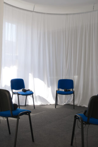 Vier blaue Stühle stehen kreisförmig auf einem grauen Teppich, auf jeder Sitzfläche liegen Kopfhörer, vor dem Hintergrund weißer Vorhänge.