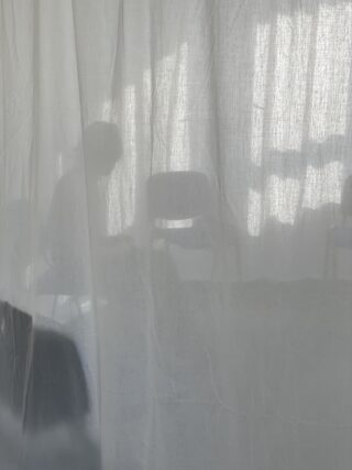 Hinter einem durchsichtigen weißen Vorhang ist die Silhouette einer auf einem Stuhl sitzenden Person zu sehen, wobei schwache Schatten und Umrisse von Stühlen sichtbar sind.