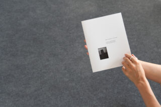 Eine Person hält vor einem schlichten grauen Hintergrund eine weiße Broschüre mit einem Schwarzweißfoto und Text auf dem Cover.