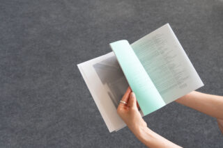 Eine Person mit einem Ring am Finger hält ein Buch mit halbtransparenten Seiten auf einer schlichten grauen Oberfläche und blättert darin.