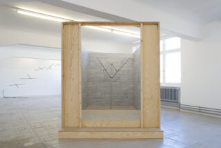 Eine Holzrahmenkonstruktion mit offener Vorderseite zeigt im Inneren eine baumartige Metallskulptur, die in einen minimalistischen Galerieraum mit weißen Wänden eingebettet ist.