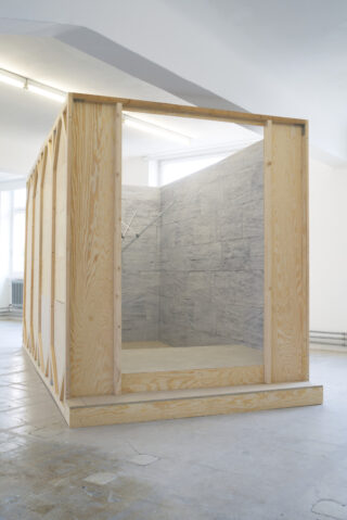 Eine große, rechteckige Holzkonstruktion mit einer offenen Seite gibt den Blick auf einen mit grauen, steinähnlichen Fliesen ausgekleideten Innenraum frei, der sich in einem schlichten, gut beleuchteten Raum befindet.