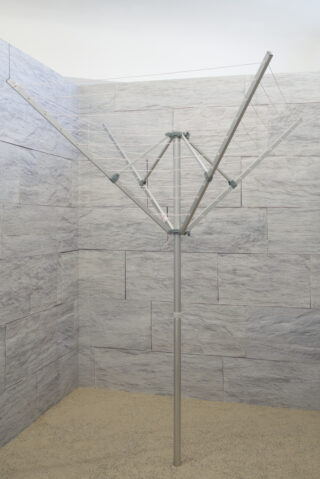 Eine Wäschespinne aus Metall ist in einem ummauerten, gefliesten Bereich mit Sandboden installiert. Die Wäscheleinenarme sind ausgefahren, aber leer.