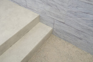 Zwei Betonstufen treffen auf eine Wand mit grauen Steinfliesen, neben der sich ein Stück groben, hellen Kieses befindet.
