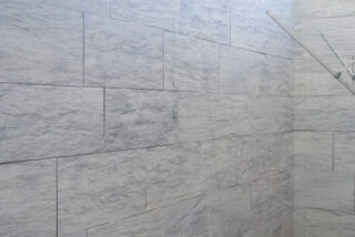 Hellgraue Steinwand mit rechteckigen Fliesen; in der oberen rechten Ecke ist ein Teil eines Wäscheständers aus Metall sichtbar.