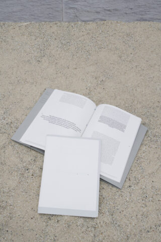 Zwei offene Bücher mit grauem Einband liegen auf einer sandigen, strukturierten Oberfläche; eines zeigt gedruckten Text, während das andere größtenteils leer ist.