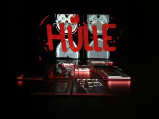 Ein dunkler Raum mit beleuchteten medizinischen Scans an der Wand, über denen in Rot das Wort „HÜLLE“ liegt, und auf dem Boden ausgestellten elektronischen Leiterplatten.