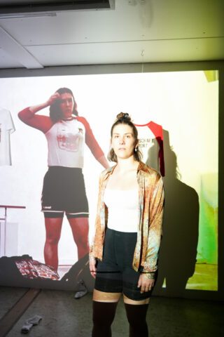 Eine Frau steht vor einem projizierten Bild von sich selbst, auf dem sie ein T-Shirt und Shorts trägt, wodurch ein vielschichtiger visueller Effekt entsteht.