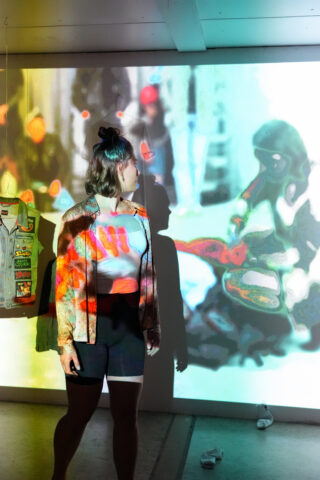 Eine Person steht vor einer bunten, abstrakten Projektion an einer Wand. Ihre Kleidung und ihr Schatten werden von der Projektion beleuchtet. Links davon hängt eine Weste.