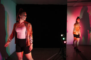 Eine Frau in Sportkleidung steht in einem schwach beleuchteten Raum mit bunten Lichtern und blickt nach vorne, während ihr Spiegelbild in einem Spiegel auf der rechten Seite erscheint.