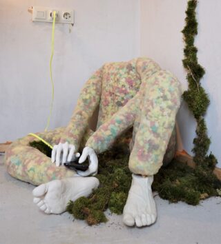 Eine kopflose, menschenähnliche Gestalt in einem durchsichtigen Anzug voller bunter Gegenstände sitzt auf dem Boden, hält einen Gamecontroller in der Hand, ist von Moos umgeben und an eine Steckdose angeschlossen.