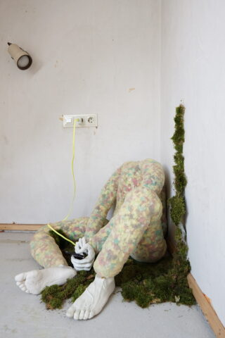 Eine kopflose, abstrakte menschliche Figur sitzt in einer Ecke, ein gelbes Kabel ist in die Wand eingesteckt, umgeben von verstreutem Moos auf dem Boden und an der Wand.