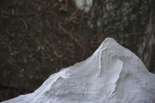 Eine Nahaufnahme einer weißen, strukturierten Skulptur in Form eines Berggipfels mit einem unscharfen natürlichen Hintergrund aus Zweigen und Blättern.