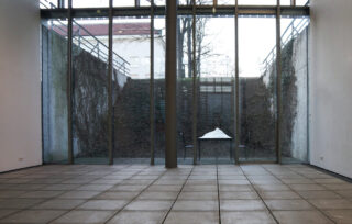 Durch eine Glaswand gibt sich der Blick auf einen Innenhof mit einem Tisch und zwei Stühlen frei. Auf dem Tisch liegt ein Haufen weißen Stoffs, umgeben von kahlen Weinreben und einem gefliesten Boden.