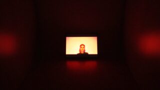 Auf einem Bildschirm wird das Gesicht einer Person in einem dunklen Raum angezeigt, wobei rotes Licht die Wände erhellt und sich auf dem Boden spiegelt.
