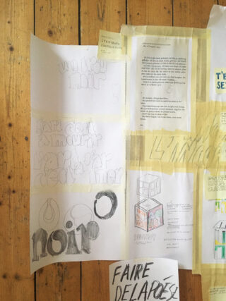 Ein Holzboden, auf dem mehrere Blätter Papier mit handschriftlichen Notizen, Skizzen und gedrucktem Text in Französisch und Englisch aufgeklebt sind.